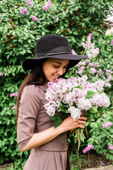 Beautiful young woman with long dark hair with a bouquet of lilacs stands in the lilac garden. Girl sniffs a lilac. Brunette in a hat with a lilac.