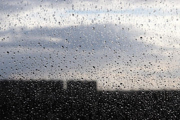Raindrops on the window glass on blurred background of city buildings and cloudy sky. Beautiful water drops, rainy weather in town