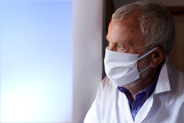 Dottore con camice bianco e mascherino guarda serio attraverso la finestra verso l'esterno