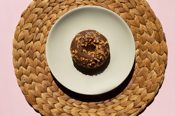 One chocolate donat decorated with caramel-flavored curls on the top at the white plate on the light brown mat, handmade from water hyacinth and clear lacquer. Pink background.Donat Day trendy concept