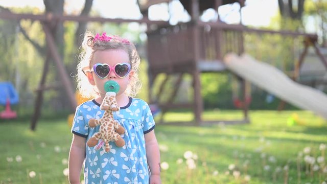 stylish little girl with pacifier and heart shaped sunglasses standing in backyard looking at camera