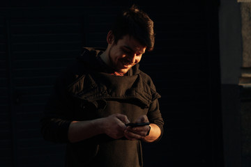 Happy smiling young man using modern smart phone while standing at crosswalk at the sunset in city center