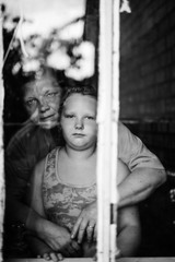 Black and white dramatic mood  portrait of grandmother hugs her granddaughter. Different generations. Old and young. Strange people looking through window. Rustic family indoor. Parents care children.