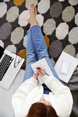 Top view woman sitting on floor indoors, using phone and laptop computer, writing notes. Studying, remote working of freelancer, creating or entertainment, relax and leisure, in cozy interior