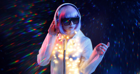 Beautiful woman with purple hair in futuristic costume and glasses over dark background. Blue and violet neon light. Portrait of young girl in modern headphones listening music. Free space for text.