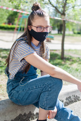 Teenage girl in protective mask sits on kerb near children's playground which is surrounded by a protective tape