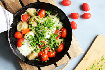 Fried eggs with cherry tomatoes, potatoes and onions in a pan on a wooden stand on the table.