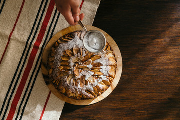 Top view of female hand using sieve to sprinkle powdered sugar over homemade natural apple pie on...