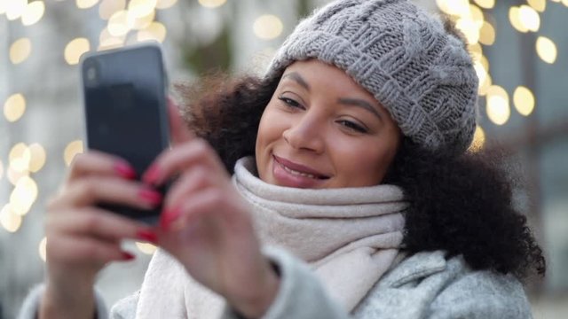 Beautiful african american woman is taking selfie using phone, standing outdoors spbi. Young curly female is looking at smartphone screen with toothy smile and making photo while having good time on