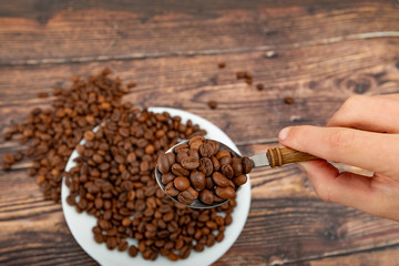 A man holds roasted coffee grains in a tablespoon on a background of a white plate and dark wood.