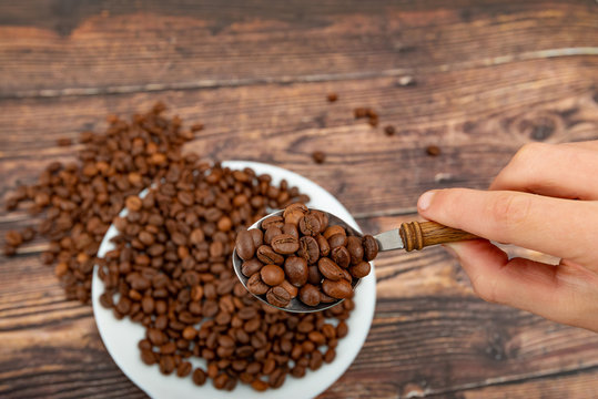 A man holds roasted coffee grains in a tablespoon on a background of a white plate and dark wood.