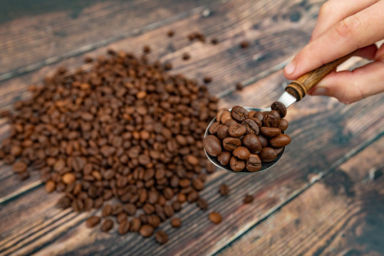 A man holds roasted coffee grains in a tablespoon on a background of a white plate and dark wood.