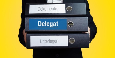 Delegat. Anwalt im Anzug trägt einen Stapel Ordner. 3 Aktenordner. Blaue Beschriftung mit Text....