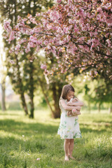 Little girl walking on the grass barefoot by the pink tree sakura in the park. Kid hug teddy bear