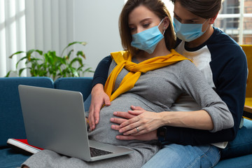 Pregnant couple in medical masks looking at the laptop screen