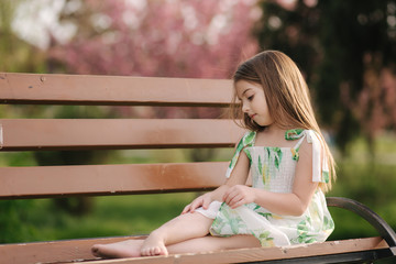 Happy female kid sits on the bench in the park. Beautiful summer dress. Fresh air. End of quarantine