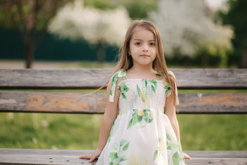 Happy female kid sits on the bench in the park. Beautiful summer dress. Fresh air. End of quarantine