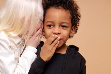 albino girl with pale skin and white hair color tells a secret in the ear of multiracial africanamerican boy. cute boy with black skin attentively listen to secret. isolated