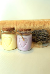 On a white background stands two cans with hearts painted on them, tied with a rope and lids from cork material. In the left bank, the cream is pastel beige, in the right lavender. Out of focus in the