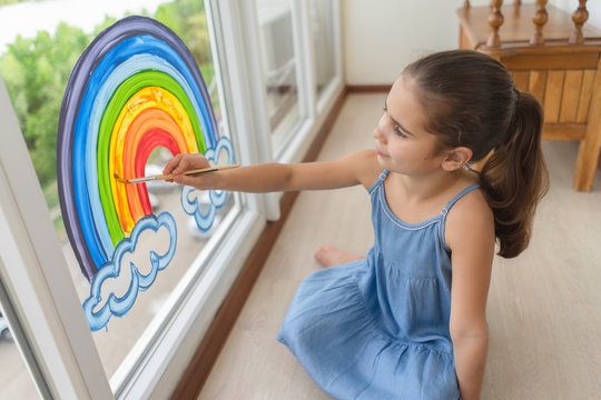 kid draws a rainbow on the window. A young girl dips a brush in paint and paints a picture of a rainbow. Close-ups on the topic of social distance and self-isolation in the concept of quarantine