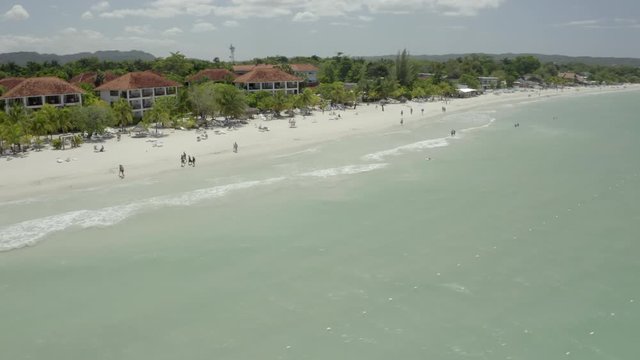 Aerial shot of people at beach near tourist resorts on sunny day, drone panning over tourists on shore near buildings during summer - Montego Bay, Jamaica