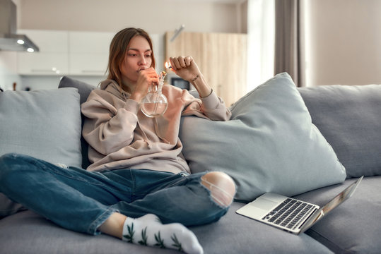 Need for Weed. Young caucasian woman sitting on the couch at home and lighting cannabis in the bowl of glass water pipe or bong. Cannabis and weed legalization concept