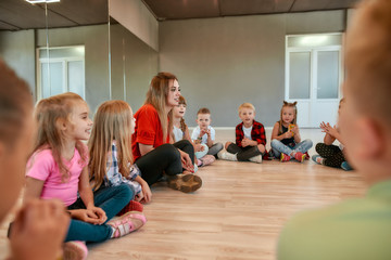 Choreography class. Young female dance teacher sitting on the floor with group of happy children in the dance studio. Little dancers sitting on the floor gathered around their female teacher