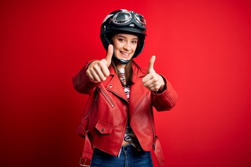 Young beautiful brunette motocyclist woman wearing motorcycle helmet and red jacket approving doing positive gesture with hand, thumbs up smiling and happy for success. Winner gesture.