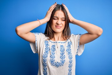 Fototapeta premium Young beautiful brunette woman wearing casual t-shirt standing over blue background suffering from headache desperate and stressed because pain and migraine. Hands on head.
