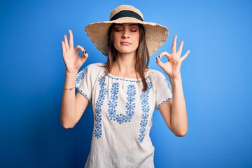 Young beautiful brunette woman wearing casual t-shirt and summer hat over blue background relax and smiling with eyes closed doing meditation gesture with fingers. Yoga concept.