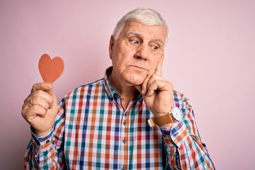 Senior handsome hoary romantic man holding red paper heart shape over pink background serious face thinking about question, very confused idea