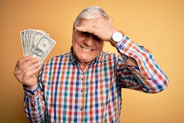 Senior handsome hoary man holding bunch of dollars banknotes over yellow background stressed with hand on head, shocked with shame and surprise face, angry and frustrated. Fear and upset for mistake.