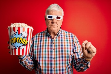 Senior hoary man watching film using 3d glasses eating popcorn over red background with a confident expression on smart face thinking serious