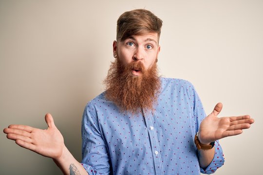 Handsome Irish redhead business man with beard standing over isolated background clueless and confused expression with arms and hands raised. Doubt concept.