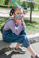 A little girl in a protective mask sits on the curb near the playground, which is surrounded by a protective tape