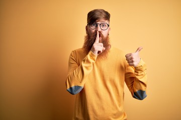 Handsome Irish redhead man with beard wearing glasses over yellow isolated background asking to be quiet with finger on lips pointing with hand to the side. Silence and secret concept.