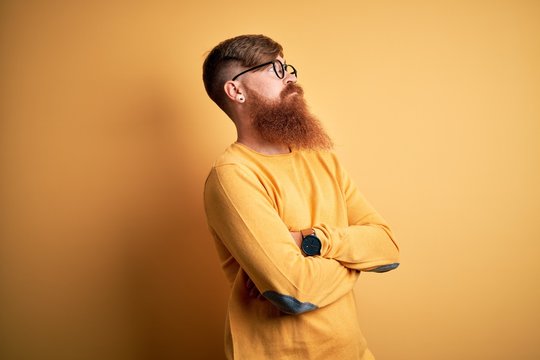 Handsome Irish redhead man with beard wearing glasses over yellow isolated background looking to the side with arms crossed convinced and confident
