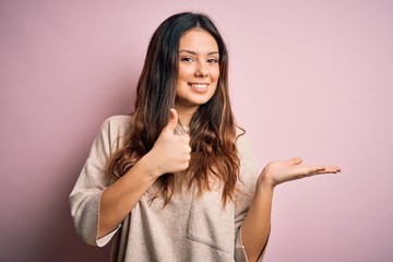 Young beautiful brunette woman wearing casual sweater standing over pink background Showing palm hand and doing ok gesture with thumbs up, smiling happy and cheerful