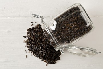 Heap of uncooked, raw, black wild rice grains in glass storage jar on white table background, flat...