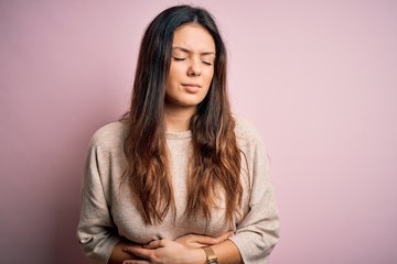 Young beautiful brunette woman wearing casual sweater standing over pink background with hand on...