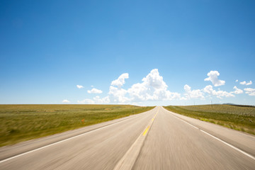 A point of view perspective of a vehicle traveling on a rural road with clouds and blue sky.