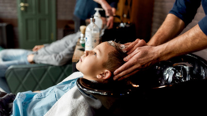 Child visiting hairdressing salon. Side view of a cute boy leaning on the sink in barbershop while male barber washing his hair © Svitlana