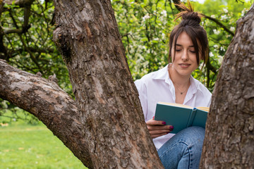 young girl in a white shirt and blue jeans sitting in nature on an apple tree reading a book. Self-isolation. Quarantine concept. Virus. Pandemic. Coronavirus