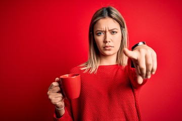 Naklejka premium Young beautiful woman drinking cup of coffee standing over isolated red background pointing with finger to the camera and to you, hand sign, positive and confident gesture from the front