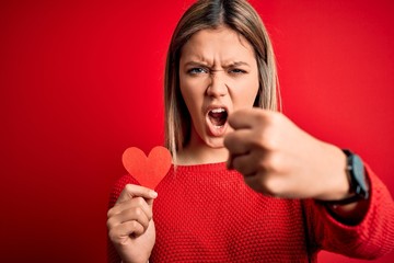 Young beautiful woman holding heart card standing over isolated red background annoyed and frustrated shouting with anger, crazy and yelling with raised hand, anger concept