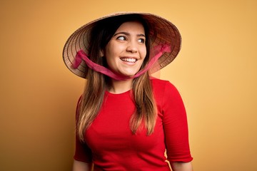 Young blonde woman wearing traditional asian rice paddy straw hat over yellow background looking away to side with smile on face, natural expression. Laughing confident.