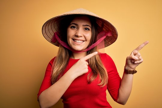 Young Blonde Woman Wearing Traditional Asian Rice Paddy Straw Hat Over Yellow Background Smiling And Looking At The Camera Pointing With Two Hands And Fingers To The Side.