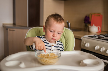 A small boy is sitting at a children's table and eating spaghetti from a plate. The baby learns to eat with a fork on its own.