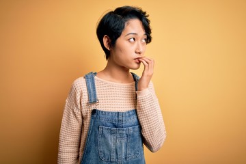 Young beautiful asian girl wearing casual denim overalls over isolated yellow background looking stressed and nervous with hands on mouth biting nails. Anxiety problem.