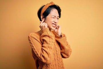 Young beautiful asian girl wearing casual sweater and diadem standing over yellow background covering ears with fingers with annoyed expression for the noise of loud music. Deaf concept.